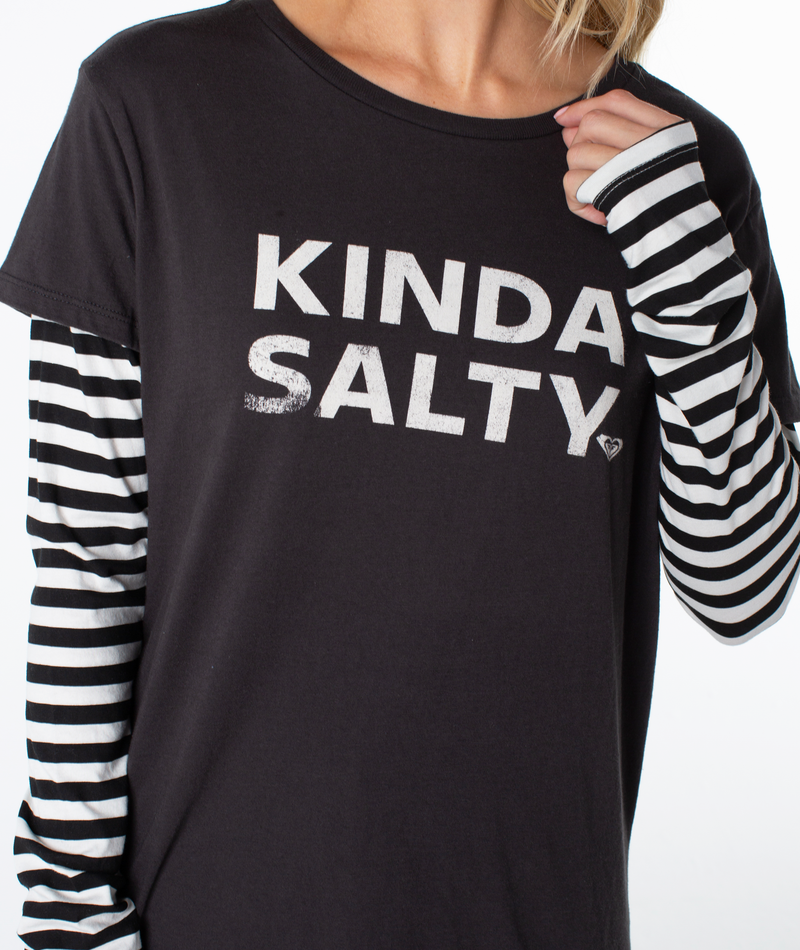 A person wears the Roxy Kinda Salty Two-fer Top—a black sporty graphic tee layered over black and white striped sleeves—for a modern coastal vibe.