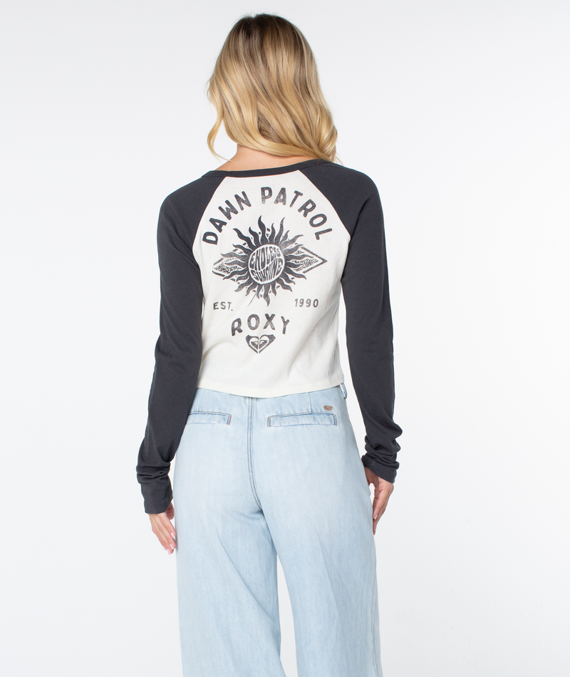 A person with long blonde hair, in light blue jeans and a white and black Roxy Sun Surfboard Boyfriend Raglan Long Sleeve, stands facing away from the camera.