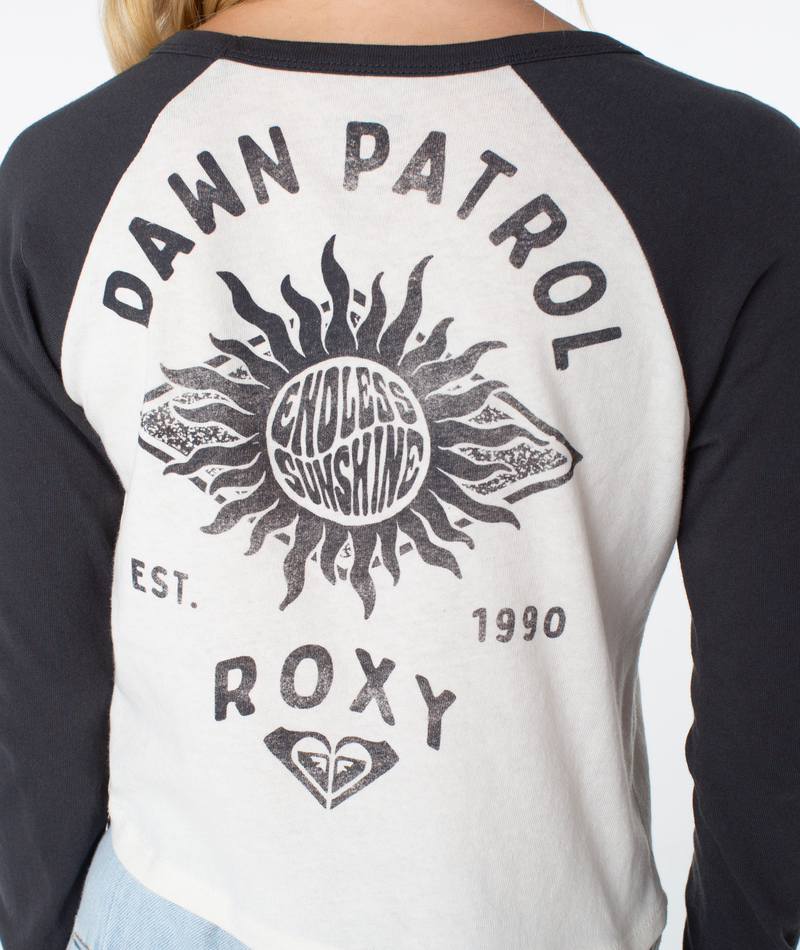 A person wears the Sun Surfboard Boyfriend Raglan Long Sleeve by Roxy, featuring a baseball-style white and black design with DAWN PATROL, ENDLESS SUNSHINE, graphics of a sun and waves, and EST. 1990 on the back.