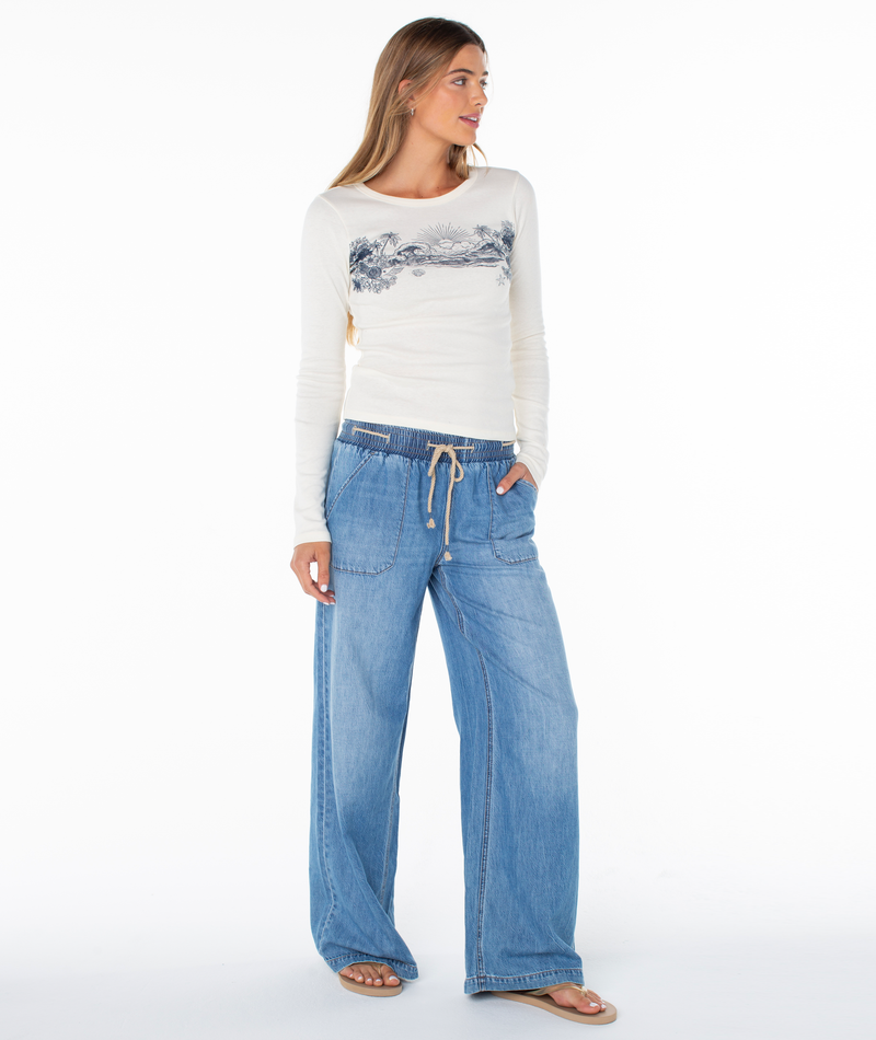 A person wearing a cream Roxy On Holiday Baby Rib Long Sleeve Top with a coastal cool graphic print and wide-leg blue jeans with a drawstring waist.