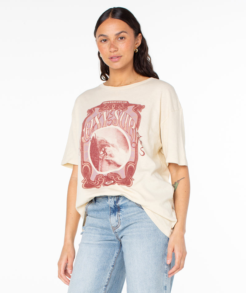 A woman wears the Roxy The Barrel Tee, an oversized cream-colored t-shirt featuring a vintage-style graphic of a surfer inside a red ornate frame.