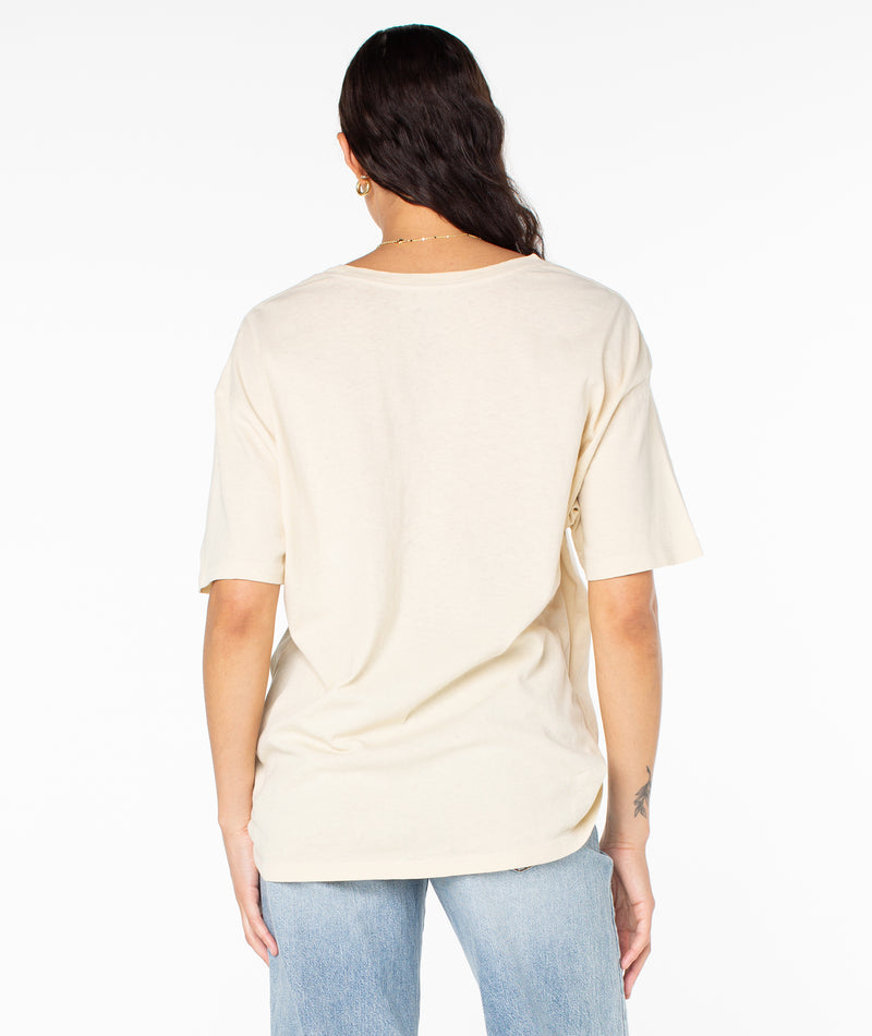 Back view of a person wearing a beige short-sleeved Roxy The Barrel Tee and blue jeans.