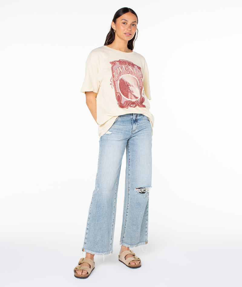 A model wears a cream short-sleeved Roxy The Barrel Tee with a maroon print, paired with distressed light-wash wide-leg jeans and tan slide sandals.