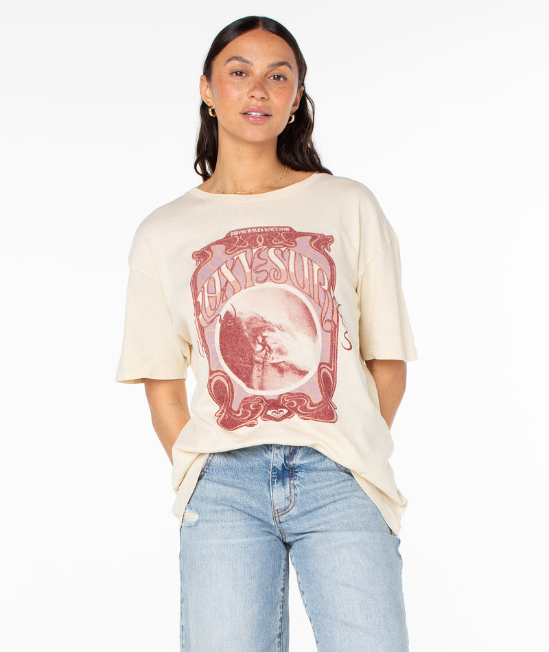 A person wears a parchment The Barrel Tee with short sleeves, featuring a vintage-style graphic of a surfer and the text Riding Waves Since 1990.