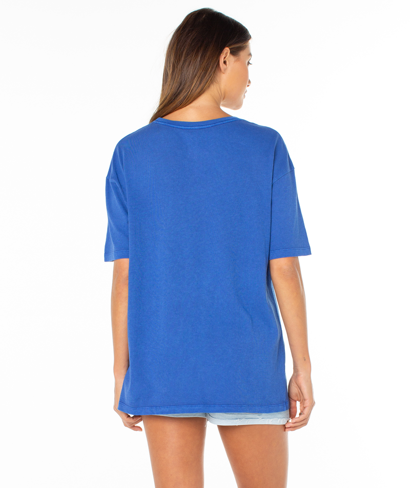A woman with long brown hair stands with her back to the camera, wearing the Roxy In Bloom Tee and light denim shorts against a plain white background.