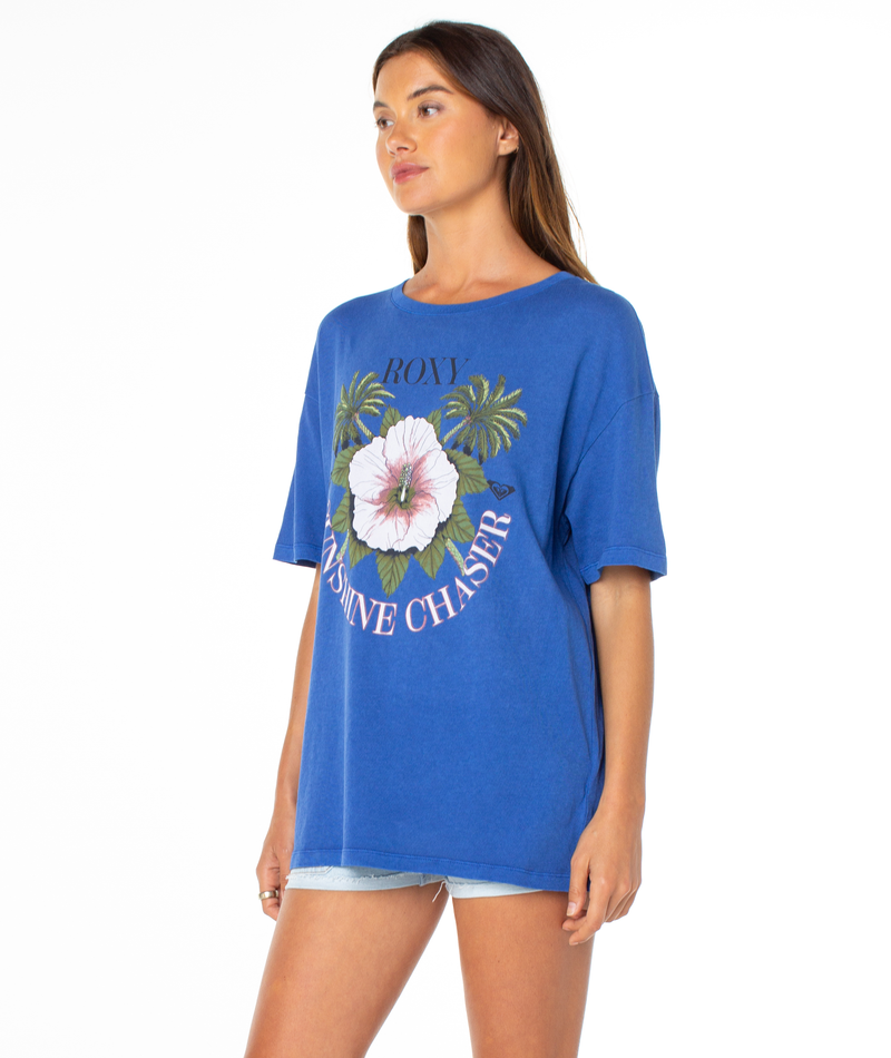 A woman models the Roxy In Bloom Tee—a loose blue shirt with a hibiscus and palm leaf graphic and “Roxy Sunshine Chaser”—paired with light denim shorts, standing in front of a plain white background.
