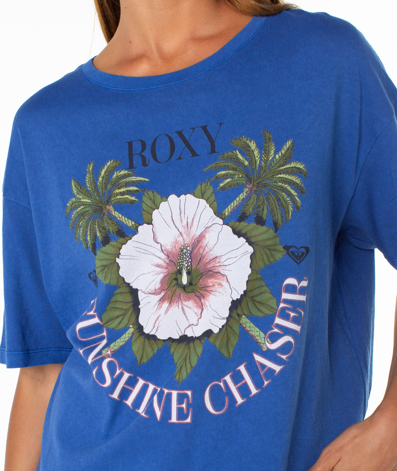 A person wearing Roxy’s In Bloom Tee—an oversized top featuring a bold white hibiscus, green palm trees, and “ROXY SUNSHINE CHASER” on the front—for a perfect beachy-chic look on sunny days.