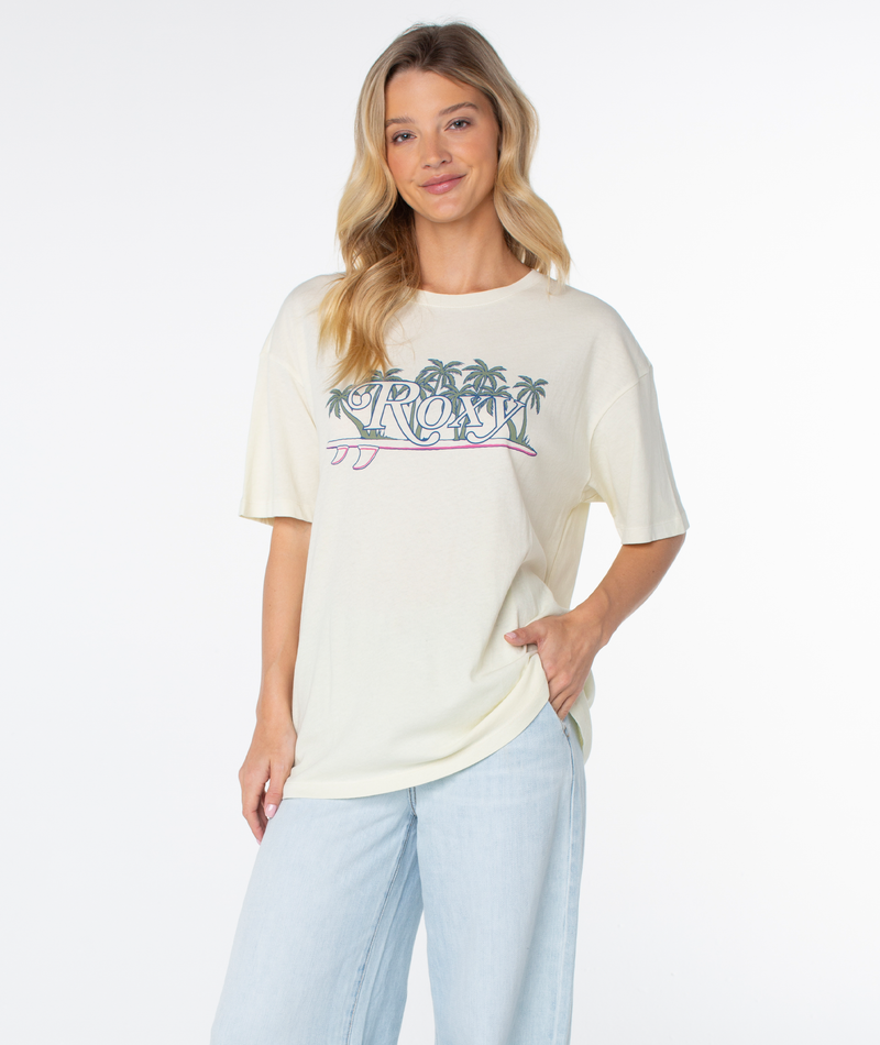 A woman wears a Roxy Sunny State Tee, an oversized, off-white short-sleeve shirt featuring a beachy-chic palm tree graphic, paired with light wash jeans.
