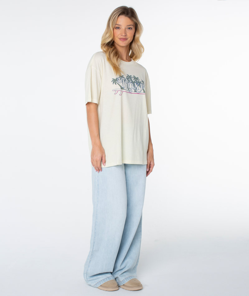 A person wears a cream Roxy Sunny State Tee with a palm tree and surfboard design, paired with wide-leg light blue jeans.