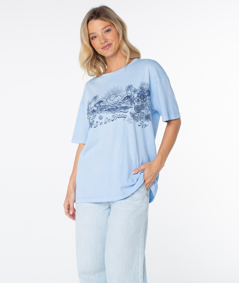 A woman wearing the light blue On Holiday Top, an oversized crew neck t-shirt featuring a beachy-chic graphic of a beach scene, palm trees, and the text I’m On Holiday.