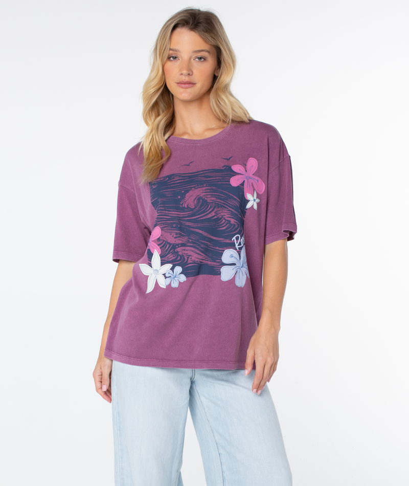 Woman wearing a Roxy In The Waves Top, an oversized, faded purple crewneck t-shirt with a beachy-chic graphic of ocean waves and hibiscus flowers on the front.