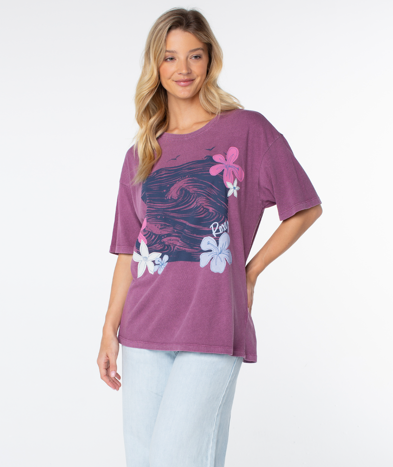 A person wears the Roxy In The Waves Top, an oversized purple t-shirt featuring a large graphic of a dark blue ocean wave, floral accents, and script text.