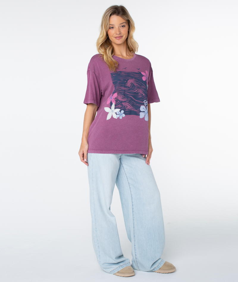 A woman wearing a purple Roxy In The Waves Top with a beachy-chic graphic of ocean waves and flowers, paired with light-wash wide-leg jeans.