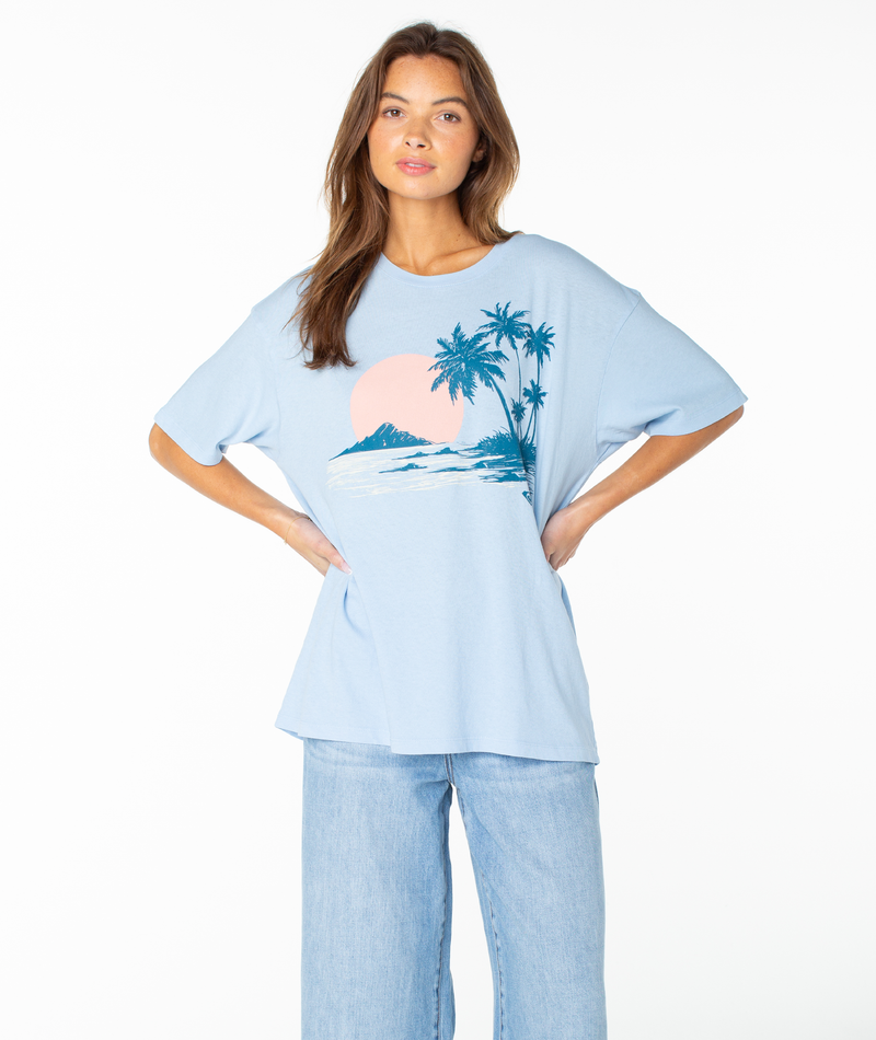 A woman stands with hands on hips against a plain white background, wearing the Roxy Classic Scenic Oversized Tee in Cerulean Classic Scenic—a light blue graphic top featuring a tropical island, palm trees, and sunset design—paired with light blue jeans.