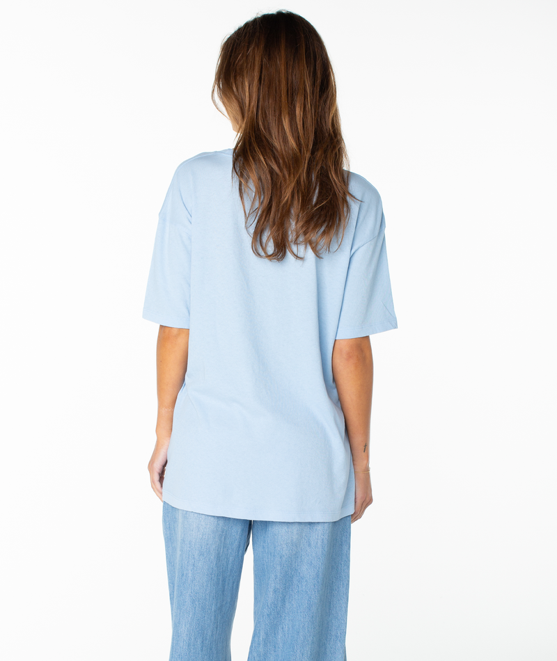 A person with long brown hair, wearing the Roxy Classic Scenic Oversized Tee in light blue and blue jeans, stands facing away from the camera against a plain white background.