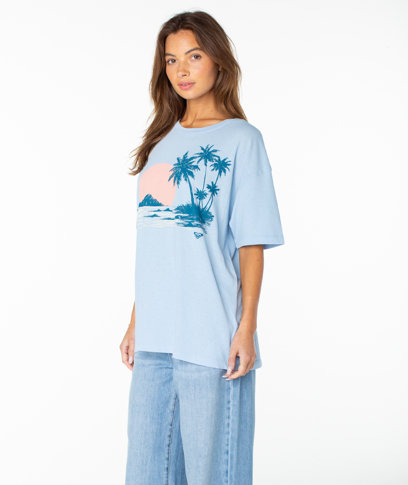 A woman wears the Roxy Classic Scenic Oversized Tee in light blue, showcasing a tropical palm tree and sunset graphic with a relaxed drop-shoulder fit, paired with light blue jeans, as she stands against a plain white background.