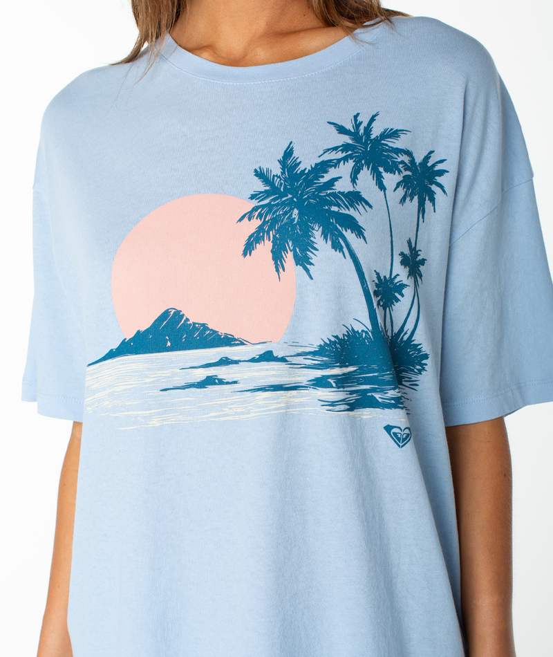 A woman wears the Roxy Classic Scenic Oversized Tee, designed with a relaxed drop shoulder and featuring a light blue graphic top decorated with a pink sun, blue palm trees, and an island scene on the front.