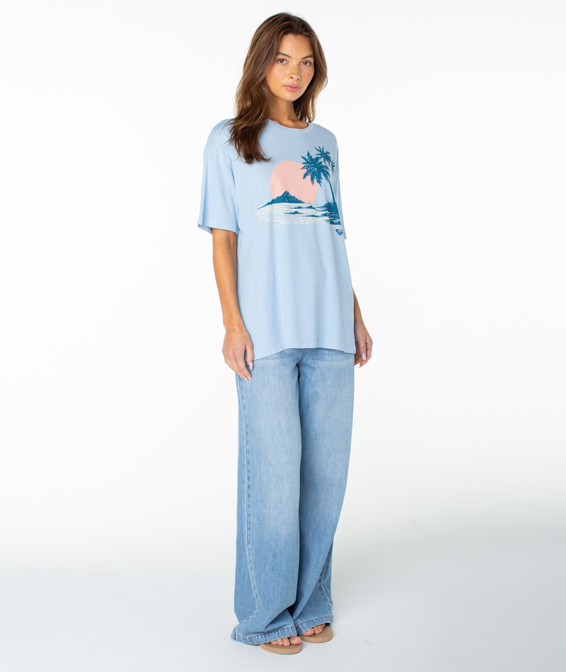 A woman stands barefoot against a plain white background, wearing the Roxy Classic Scenic Oversized Tee—a light blue graphic top with a palm tree and sunset design—paired with wide-leg blue jeans.