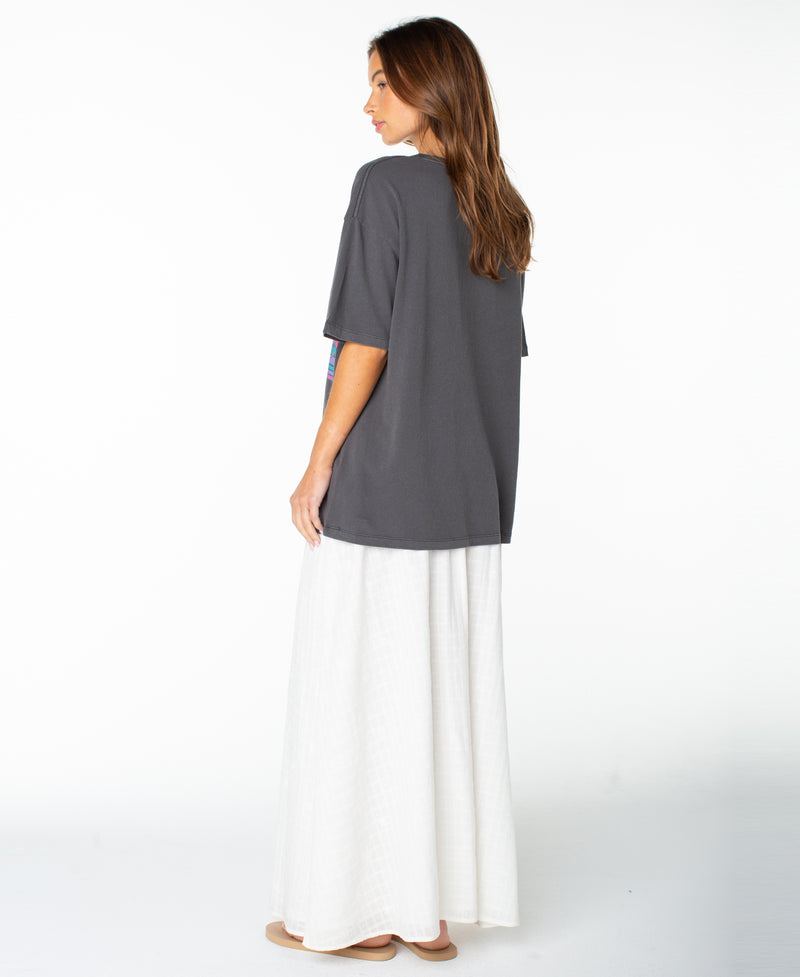 A woman stands sideways, wearing the Roxy Heatwave Oversized Tee in Phantom Heatwave paired with a long, flowing white skirt. She has long brown hair and is barefoot, set against a plain white background.
