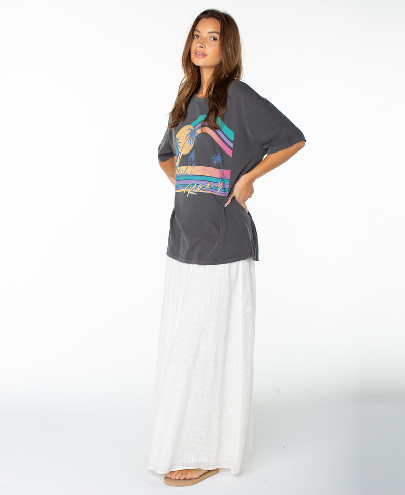 A woman stands in profile against a white background, wearing the Roxy Heatwave Oversized Tee featuring a palm tree graphic, paired with a long white skirt and sandals. She has long brown hair and her hands in the tee’s pockets.