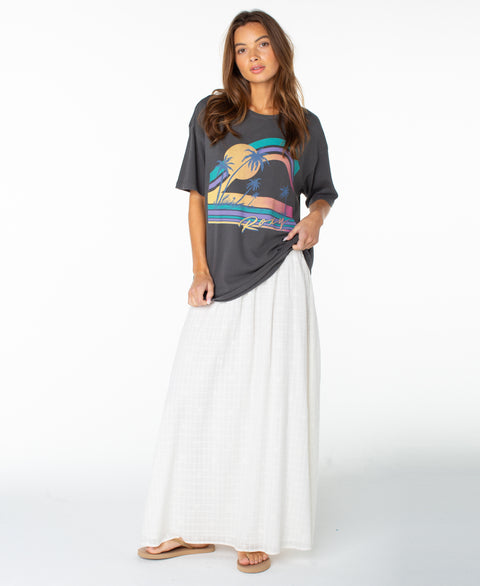 A woman stands against a plain white background wearing the Roxy Heatwave Oversized Tee with a colorful palm tree graphic, paired with a long white skirt and sandals.