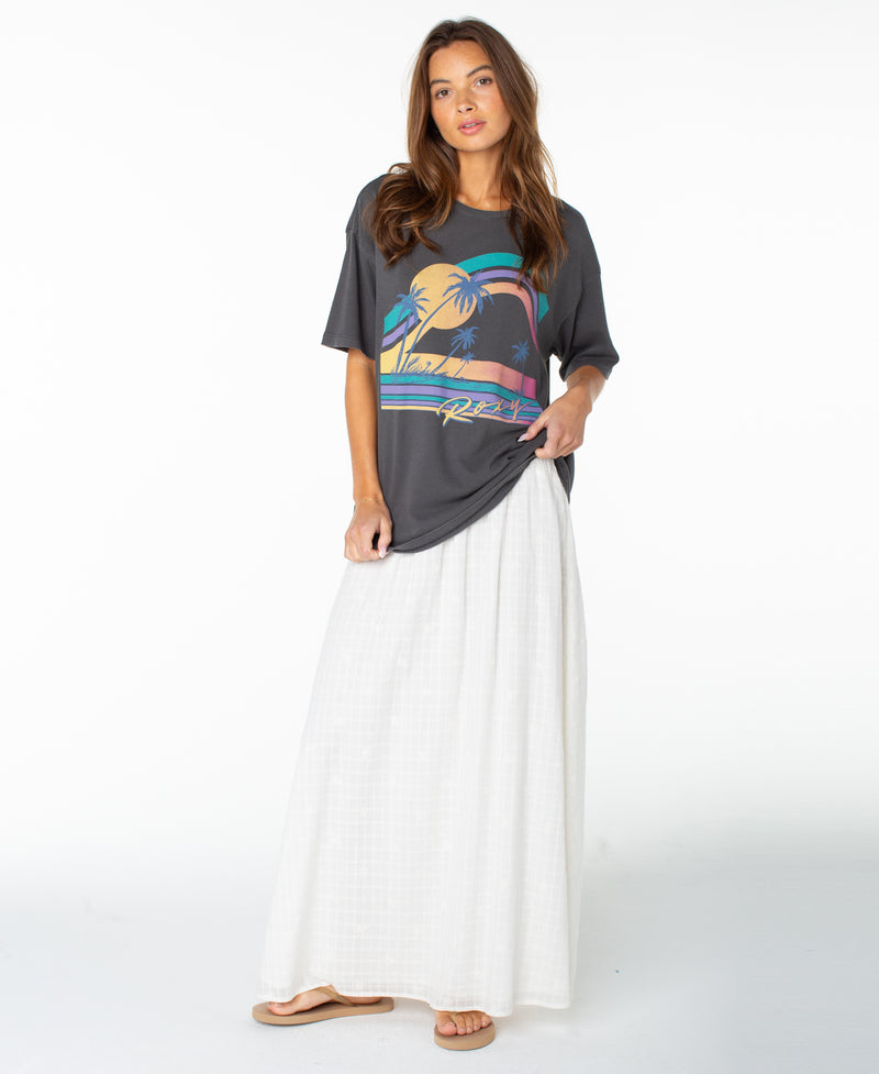 A woman stands against a plain white background wearing the Roxy Heatwave Oversized Tee with a colorful palm tree graphic, paired with a long white skirt and sandals.