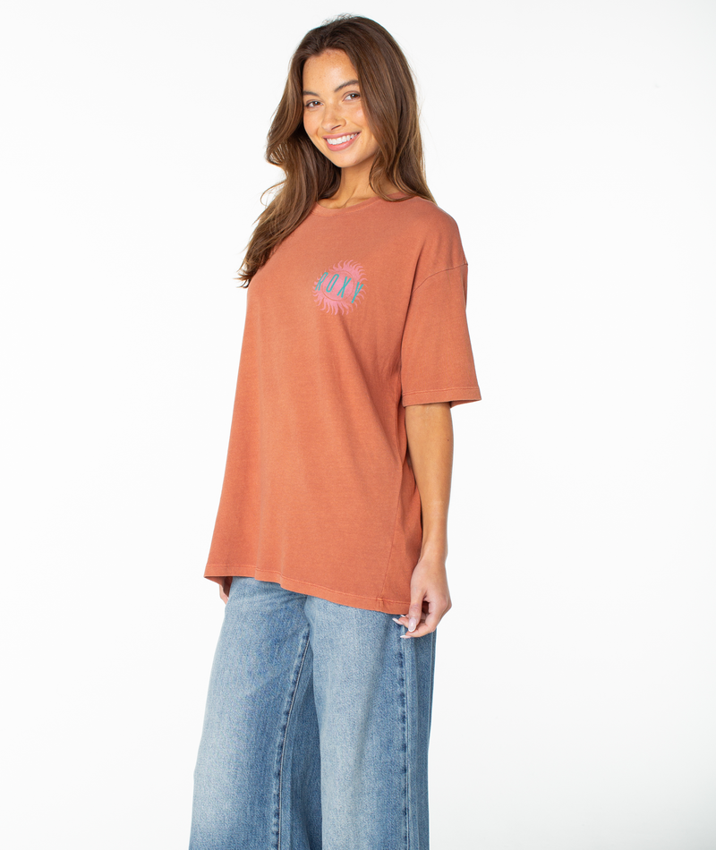 A woman with long brown hair smiles while wearing the Roxy Sun Club Oversized Tee in Chutney Sun Club, featuring a relaxed drop shoulder, small emblem, and paired with light blue wide-leg jeans as she stands against a plain white background.