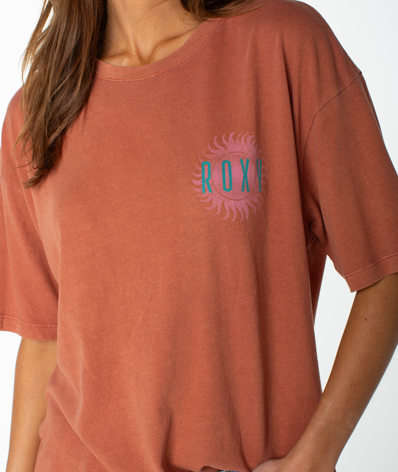 A woman wears the Sun Club Oversized Tee by Roxy, featuring a faded orange hue and a pink sunburst graphic with the ROXY logo on the chest. Only her upper body is visible against a plain white background. The relaxed drop shoulder enhances the laid-back, effortless style of this lightweight top.