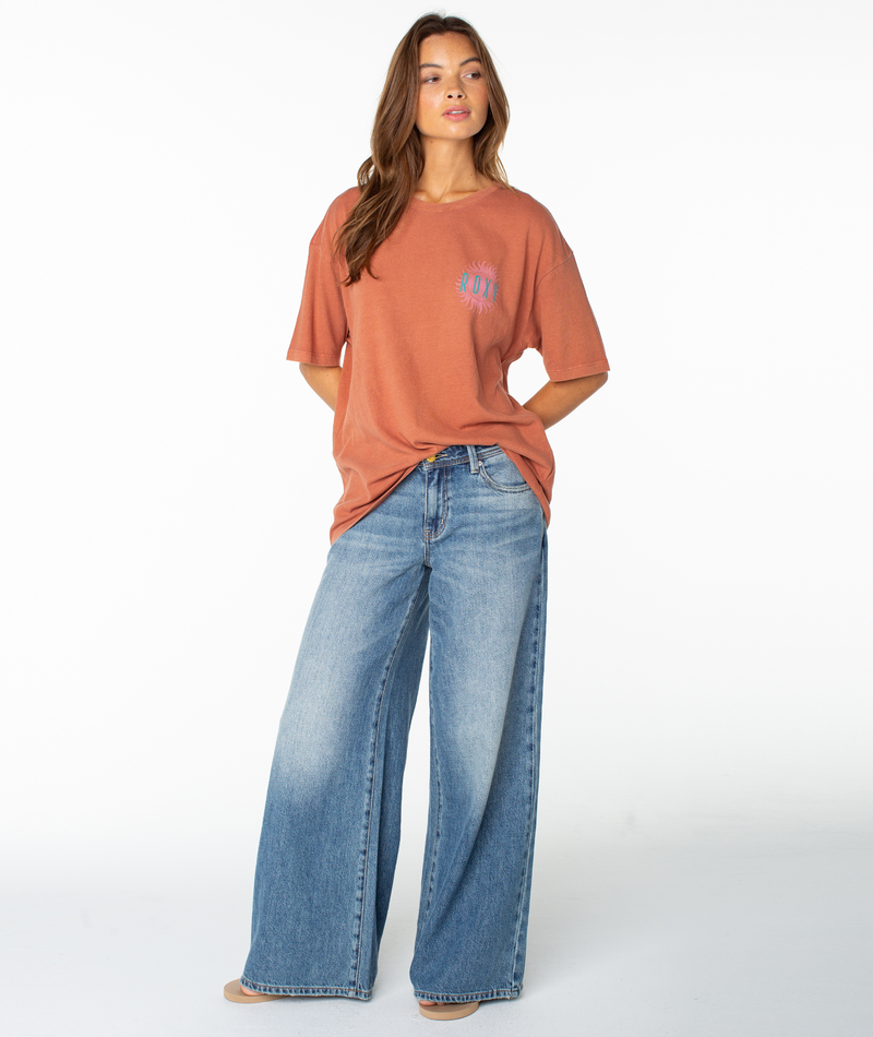 A woman stands against a white background wearing the Roxy Sun Club Oversized Tee in rust and loose-fitting blue jeans, with her hands in her pockets and looking slightly to the side.