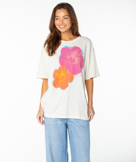 A woman smiles in the Roxy Flower Glow Oversized Tee in Cloud Dancer Flower Glow, featuring a relaxed drop shoulder design paired with light wash wide-leg jeans.