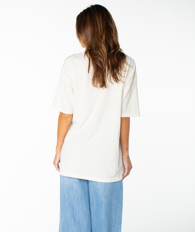 A person with long brown hair stands with their back to the camera, wearing the Roxy Flower Glow Oversized Tee with relaxed drop shoulders and wide-leg light blue jeans against a plain white background.