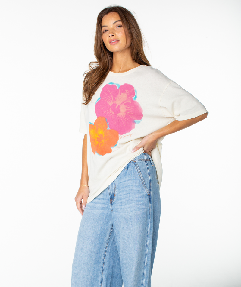 A woman with long brown hair wears the Roxy Flower Glow Oversized Tee, a relaxed white drop shoulder top with large pink and orange flowers, paired with light blue jeans. She poses confidently, one hand in her pocket and the other on her hip.