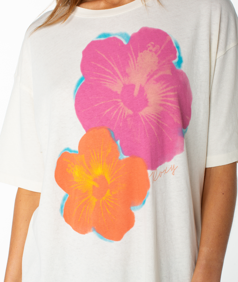 A person wears the Roxy Flower Glow Oversized Tee, a lightweight top with a relaxed drop shoulder fit featuring two large hibiscus flowers (pink and orange) with blue accents. Only the torso and part of one arm are visible.