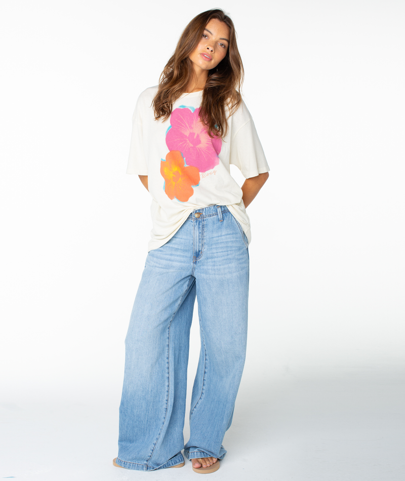 A woman with long brown hair poses in the Roxy Flower Glow Oversized Tee, featuring large pink and orange floral prints, paired with light blue wide-leg jeans and beige open-toe shoes against a white background.