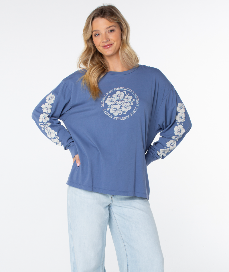 A woman smiles with hands on hips in the Roxy Vintage Hibiscus Oversized Long Sleeve Tee, featuring white floral designs—perfect for breezy coastal evenings.