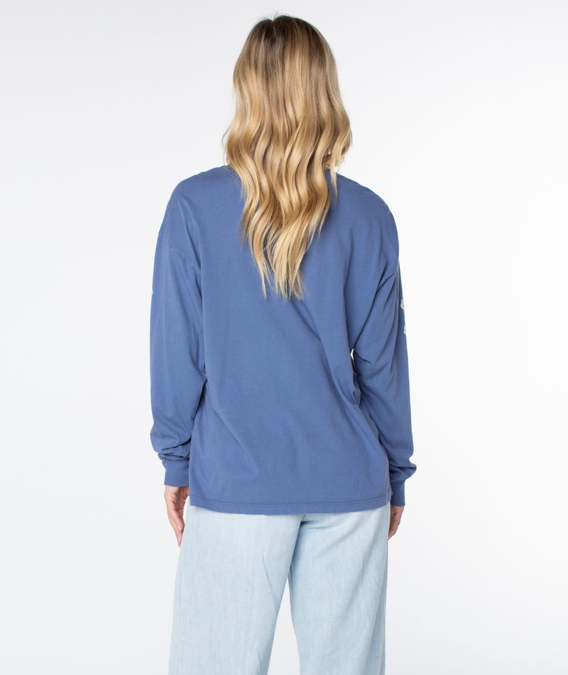 A person with long wavy blonde hair, in light blue jeans and a Roxy Vintage Hibiscus Oversized Long Sleeve Tee, faces away on a white background—a perfect coastal evening look.