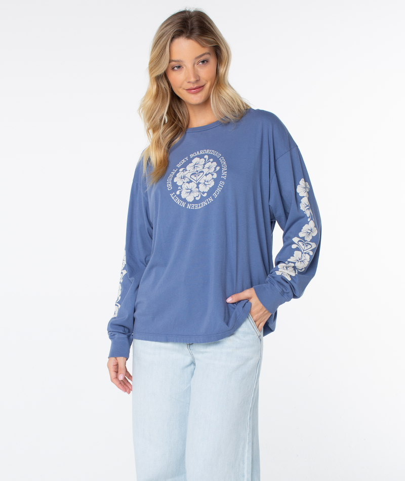 A woman with long blonde hair wears the Roxy Vintage Hibiscus Oversized Long Sleeve Tee, featuring floral designs, paired with light blue jeans against a plain white background.