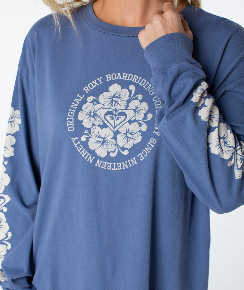 Person wears a Vintage Hibiscus Oversized Long Sleeve Tee by Roxy with white hibiscus designs on sleeves and chest. Circular text: Original Roxy Boardriding Co. Since Nineteen Ninety—perfect for coastal evenings.