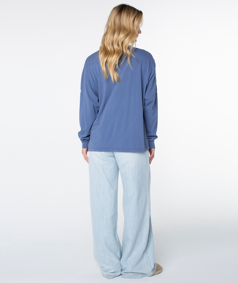 A person with long blonde hair wears the Roxy Vintage Hibiscus Oversized Long Sleeve Tee and light blue wide-leg jeans, standing with their back to a white background—ideal for cool coastal evenings.