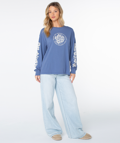 A young woman faces forward in a Roxy Vintage Hibiscus Oversized Long Sleeve Tee (Wild Wind Vintage Hibiscus) and light-wash wide-leg jeans, with wavy blonde hair and beige shoes. Plain white background.