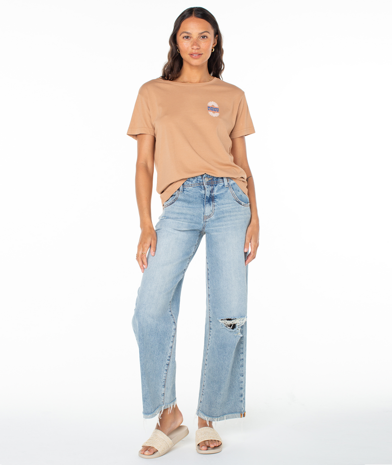 A person wearing a tan Roxy Besos Tee with a small circular chest graphic, light-wash distressed wide-leg jeans, and textured slides.