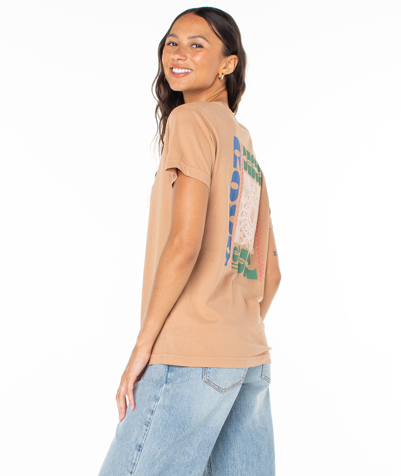 A woman wearing a tan Roxy Besos Tee with a colorful graphic on the back and light-wash jeans.