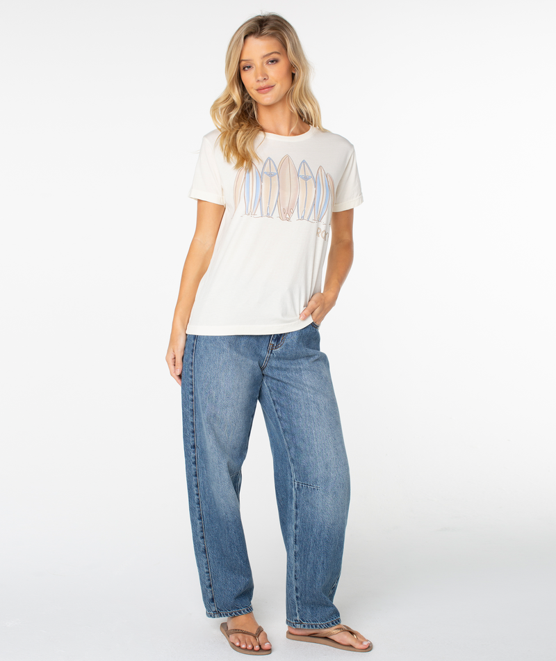 A woman wearing a cream-colored Roxy Dad Vibes Surfboard Tee featuring a surfboard graphic, paired with loose-fit blue jeans and flip-flops.