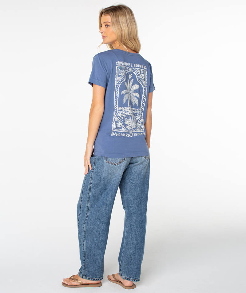 A person seen from behind wearing a blue Roxy Simple Palm Top with a white palm tree graphic and the text Paradise Bound, paired with blue jeans and sandals.