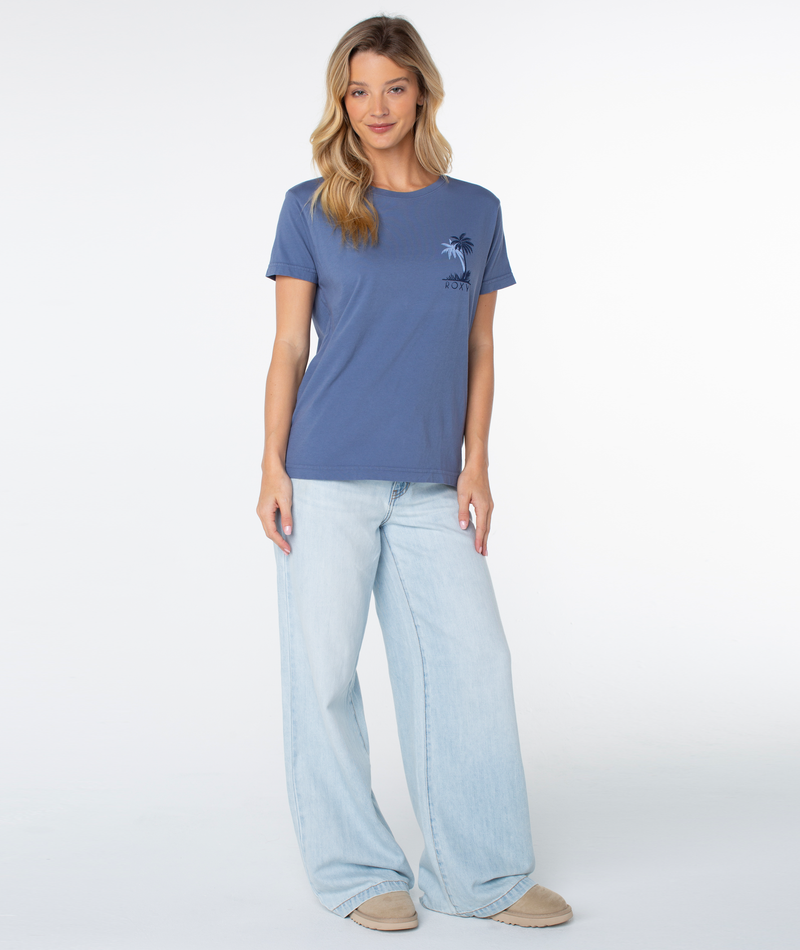 A woman wearing a Roxy Palm Geo Top in Wild Wind, a slouchy blue lounge shirt with a small palm tree graphic, paired with wide-leg light-wash jeans.
