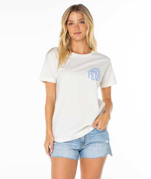 A woman with long blonde hair models the Roxy High Tides Tee—a slouchy white shirt with a small blue graphic—paired with light blue denim shorts, standing against a white background and looking at the camera with one hand in her pocket.
