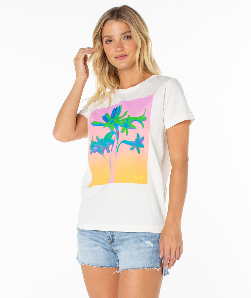 A woman with long blonde hair wears the Roxy Tuberose Tee, a white top with a colorful floral graphic, and denim shorts. She stands against a plain white background, smiling slightly with one hand touching her hair.