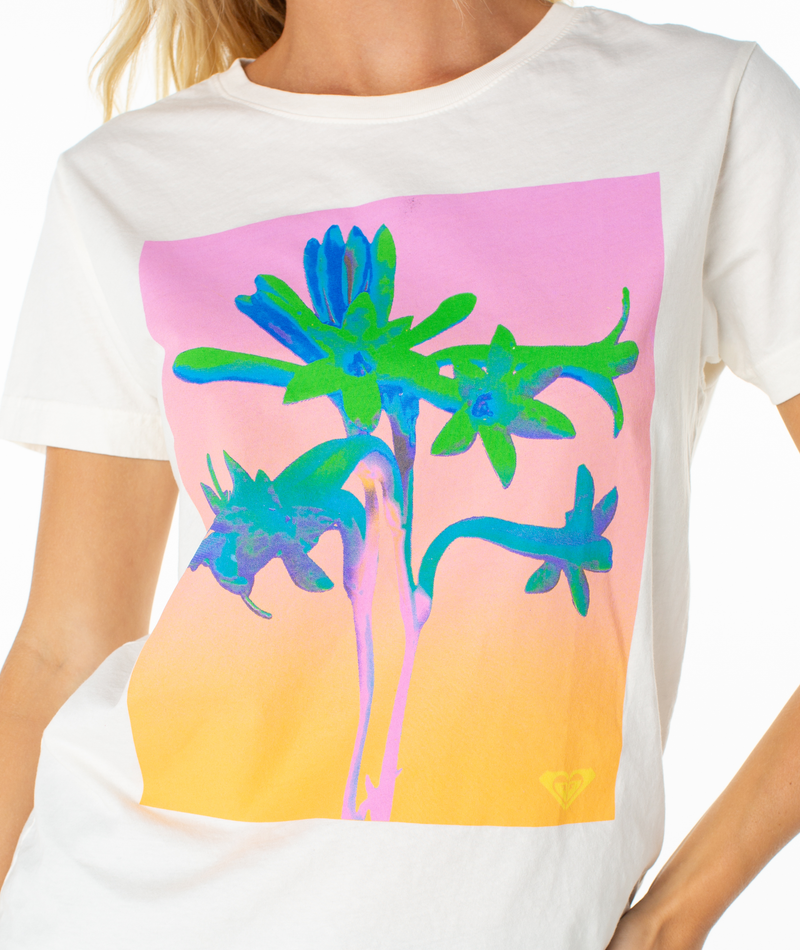 A person wears the Roxy Tuberose Tee, a white lounging top with a colorful abstract floral graphic in green, blue, and purple on a pink and orange background.