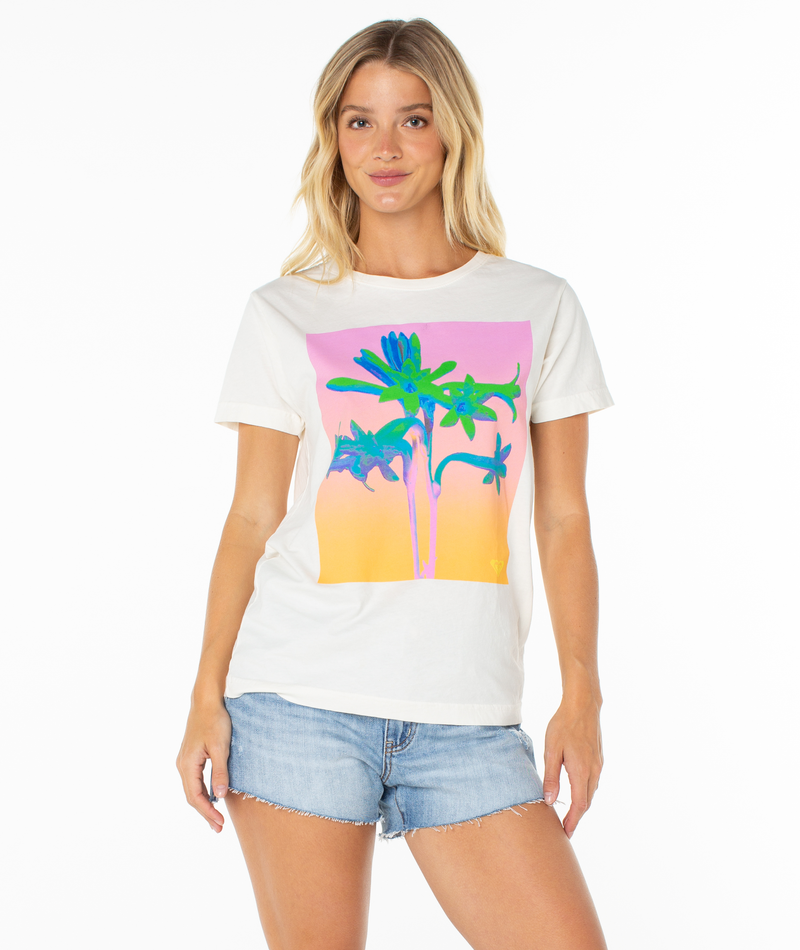 A woman with long blonde hair smiles gently as she models the Roxy Tuberose Tee—a white top with a vibrant tropical flower graphic—paired with light denim shorts, standing against a plain white background.