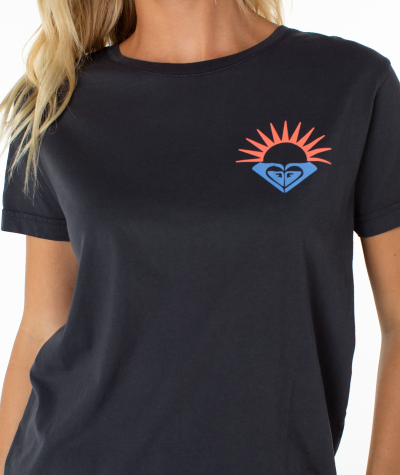 A person wearing the Roxy Surrounded By Sunshine Tee, a dark loungewear t-shirt featuring a small, colorful blue and black emblem with red-orange sun rays on the left chest.